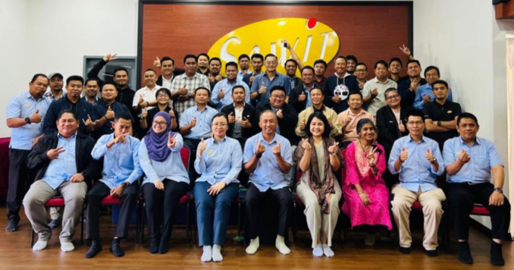 A large group of people sit smiling and posing for a photo in front of a yellow logo wall, many are giving thumbs up.